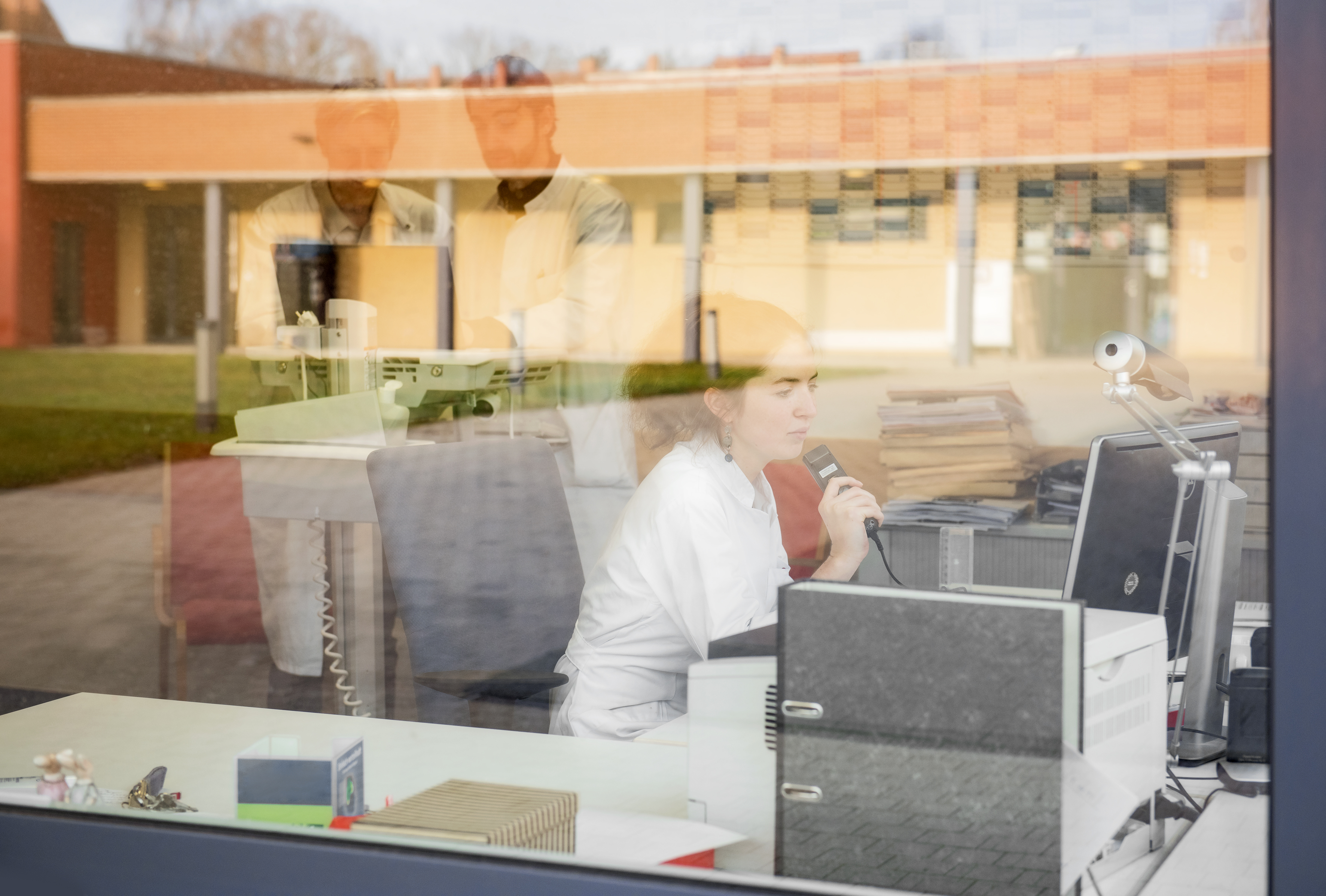 Frau sitzt an einem Computer. in der Scheibe spiegeln sich weitere Menschen