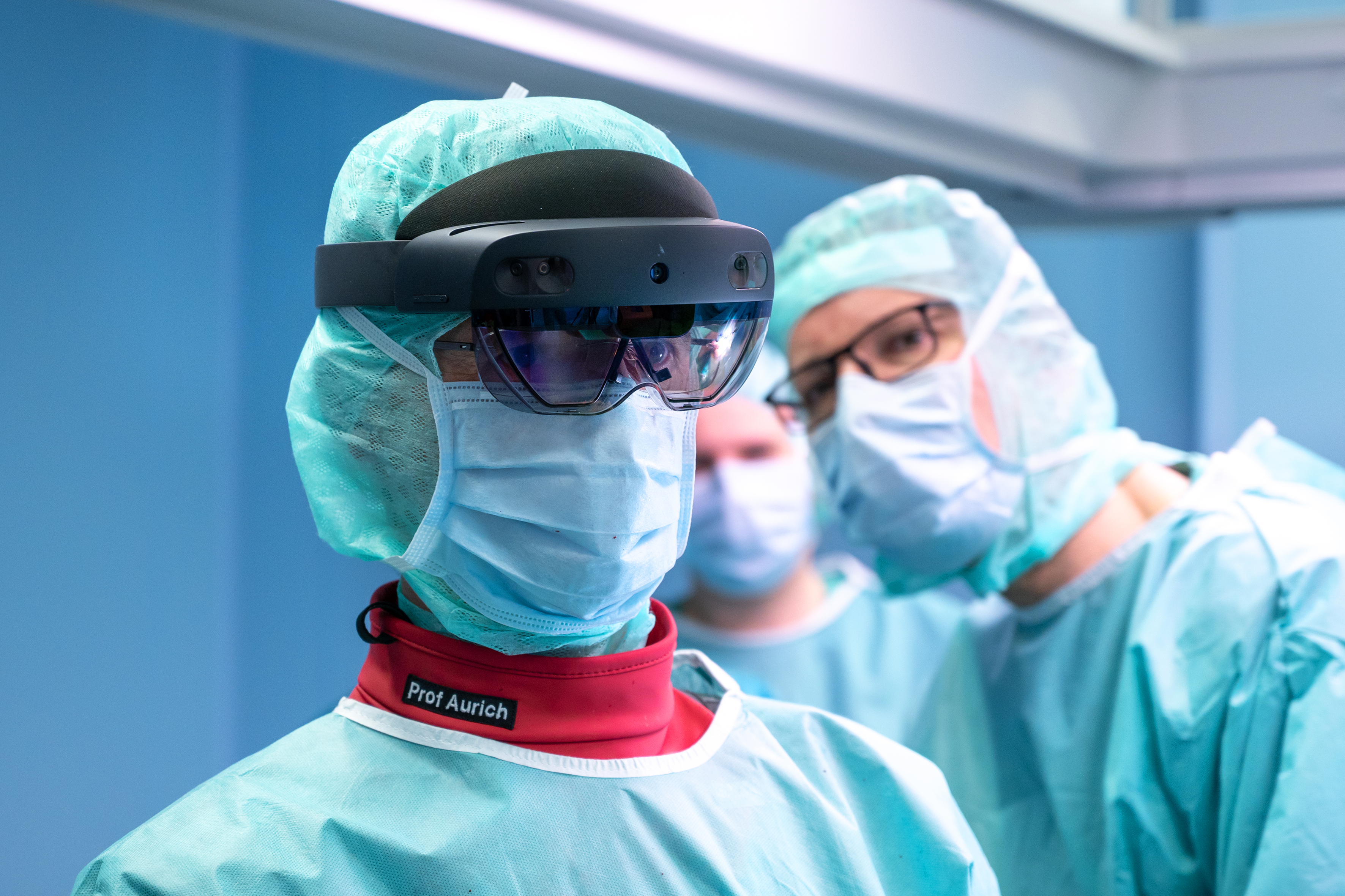 Mann im Portrait. Er trägt OP-Kleidung und eine VR-Brille. Hinter ihm stehen zwei weitere Männer in OP-Kleidung.