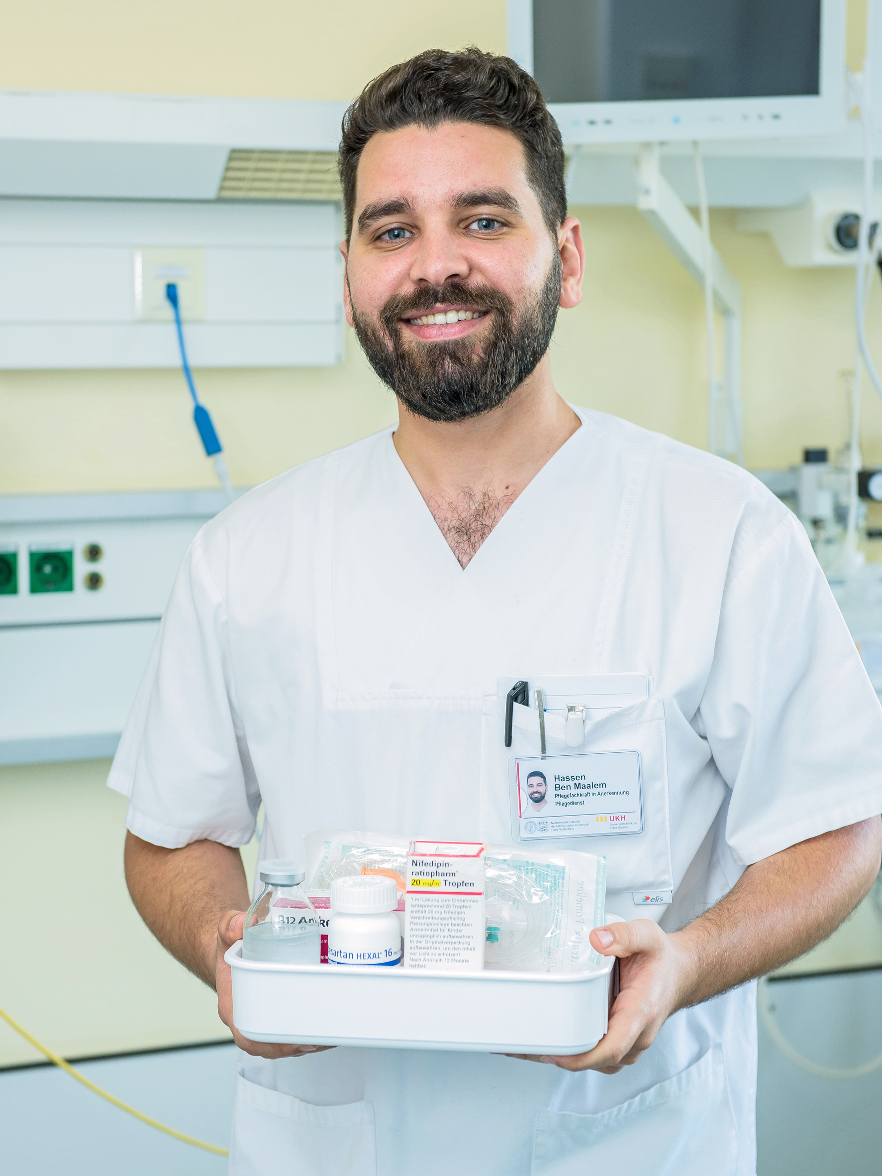 Ein junger Mann mit Bart und weißem Kassak hält ein Tablett mit medizinischem Arbeitsmaterial in den Händen. Er schaut freundlich in die Kamera.