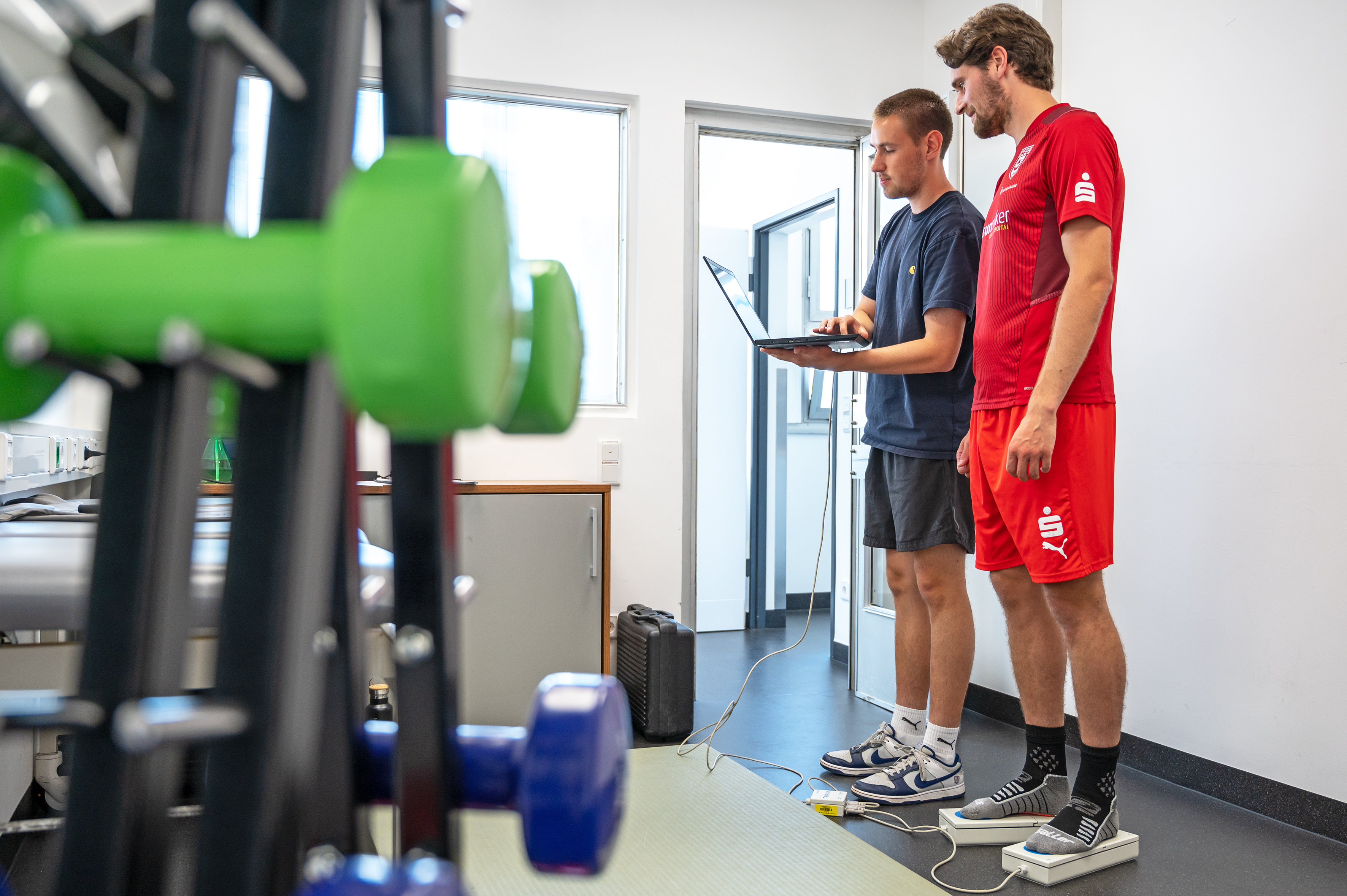 Zwei Männer stehen in einem kleinen Raum mit Gewichten im Vordergrund. Der Mann links zeigt dem Mann rechts (Jonas Nietfeld) etwas auf einem Laptop.