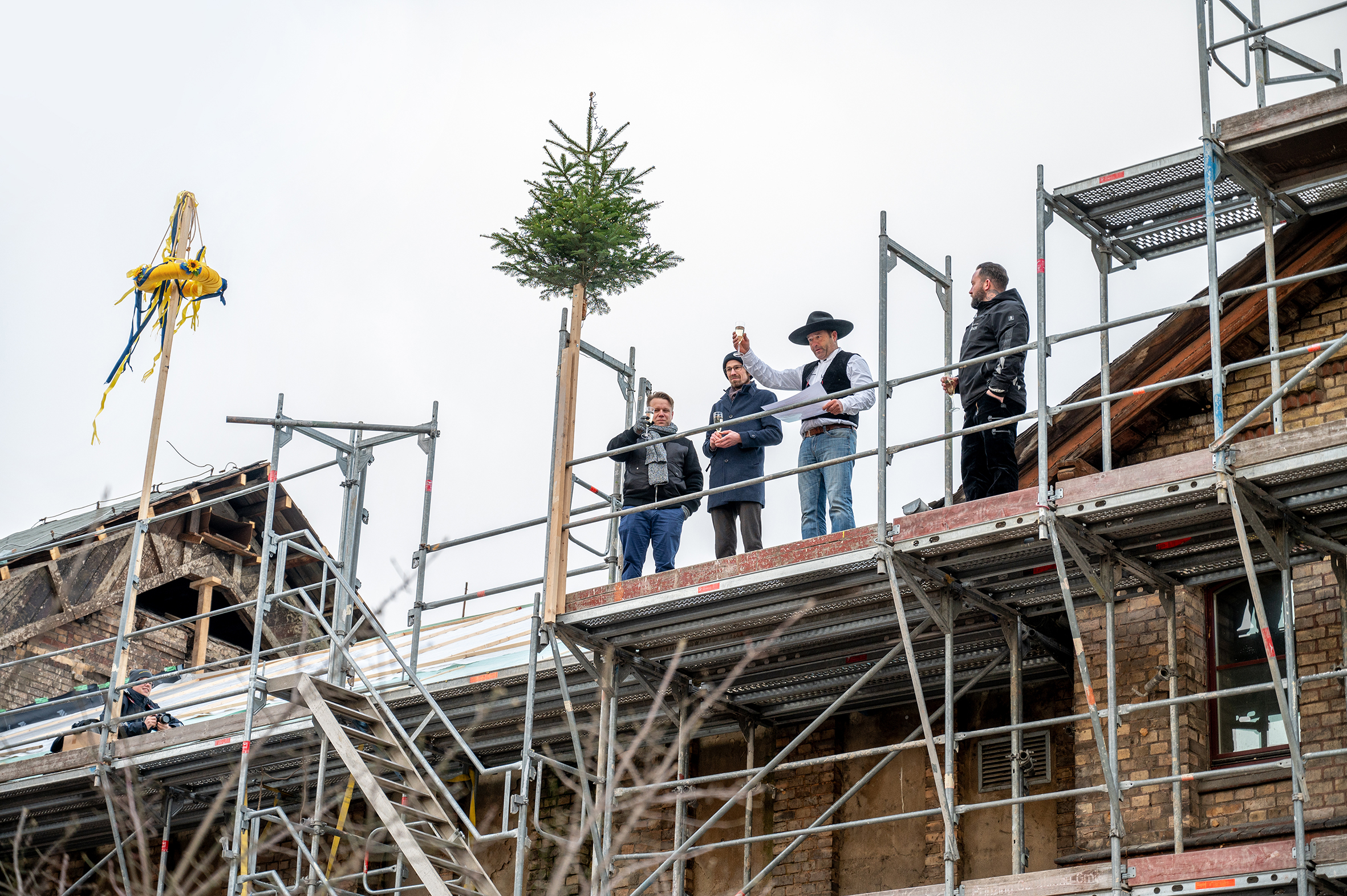 Vier Männer stehen auf einem Dachstuhl, sie haben Sektgläser in der Hand. Es ist ein traditioneller Richtkranz in gelb-blau zu sehen. Der zweite Mann von rechts trägt traditionelle Zimmermannskleidung mit Hut.