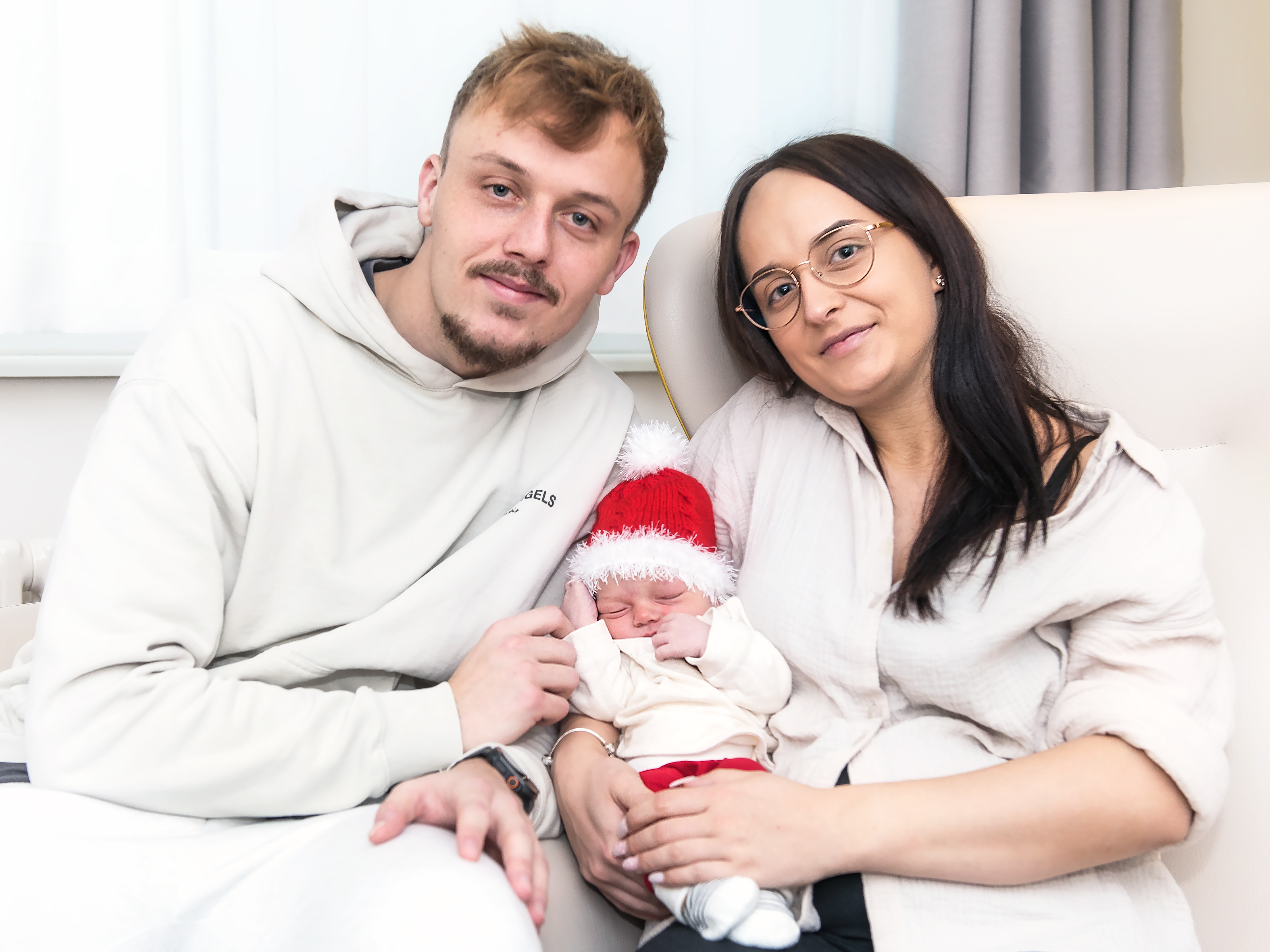 Vater und Mutter halten ein neugeborenes Baby zwischen sich. Die Eltern tragen beige Kleidung, das Baby eine Weihnachtsmütze.