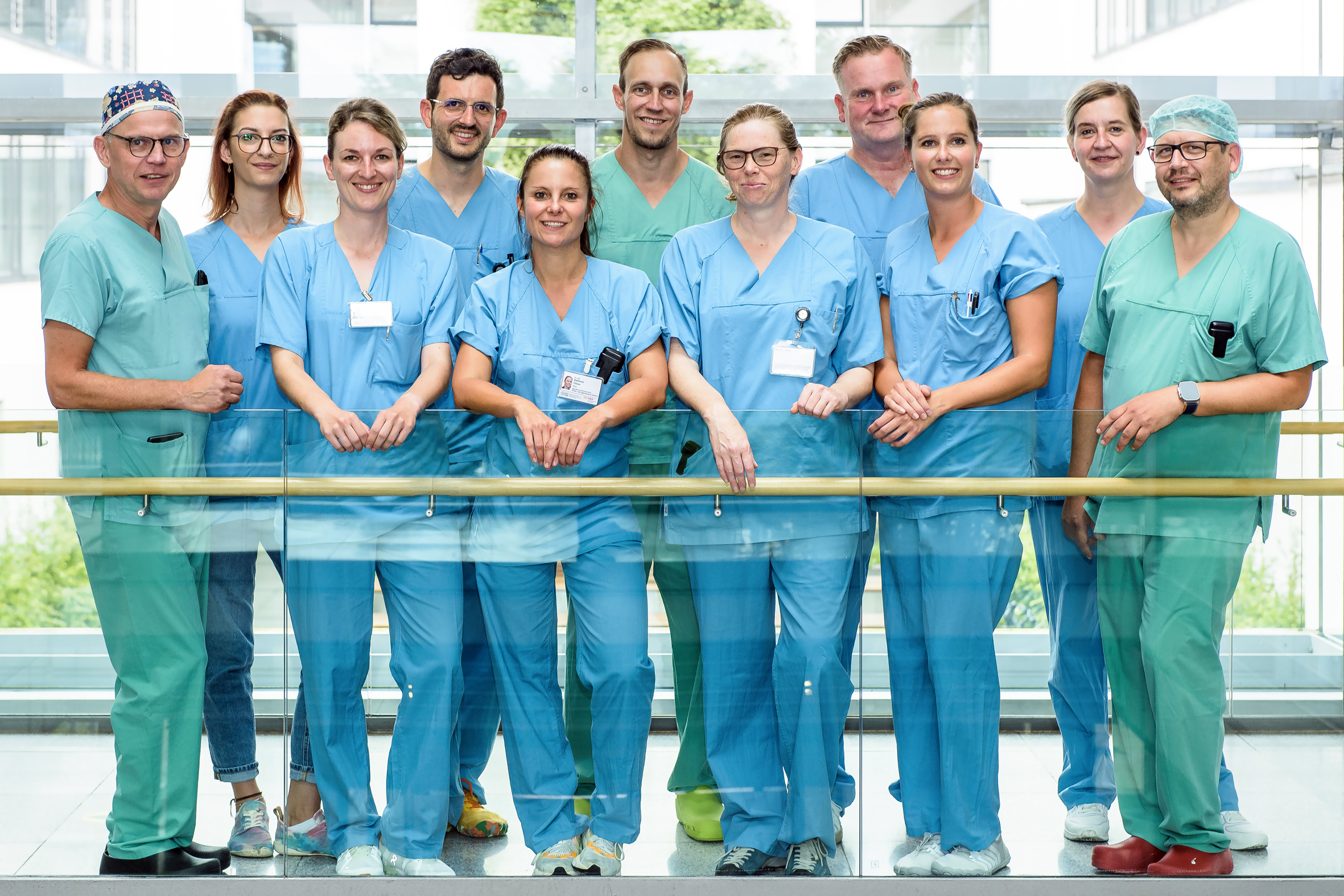 Elf Männer und Frauen in medizinischer Arbeitskleidung (blau und grün) stehen an einer Brüstung und blicken in die Kamera.