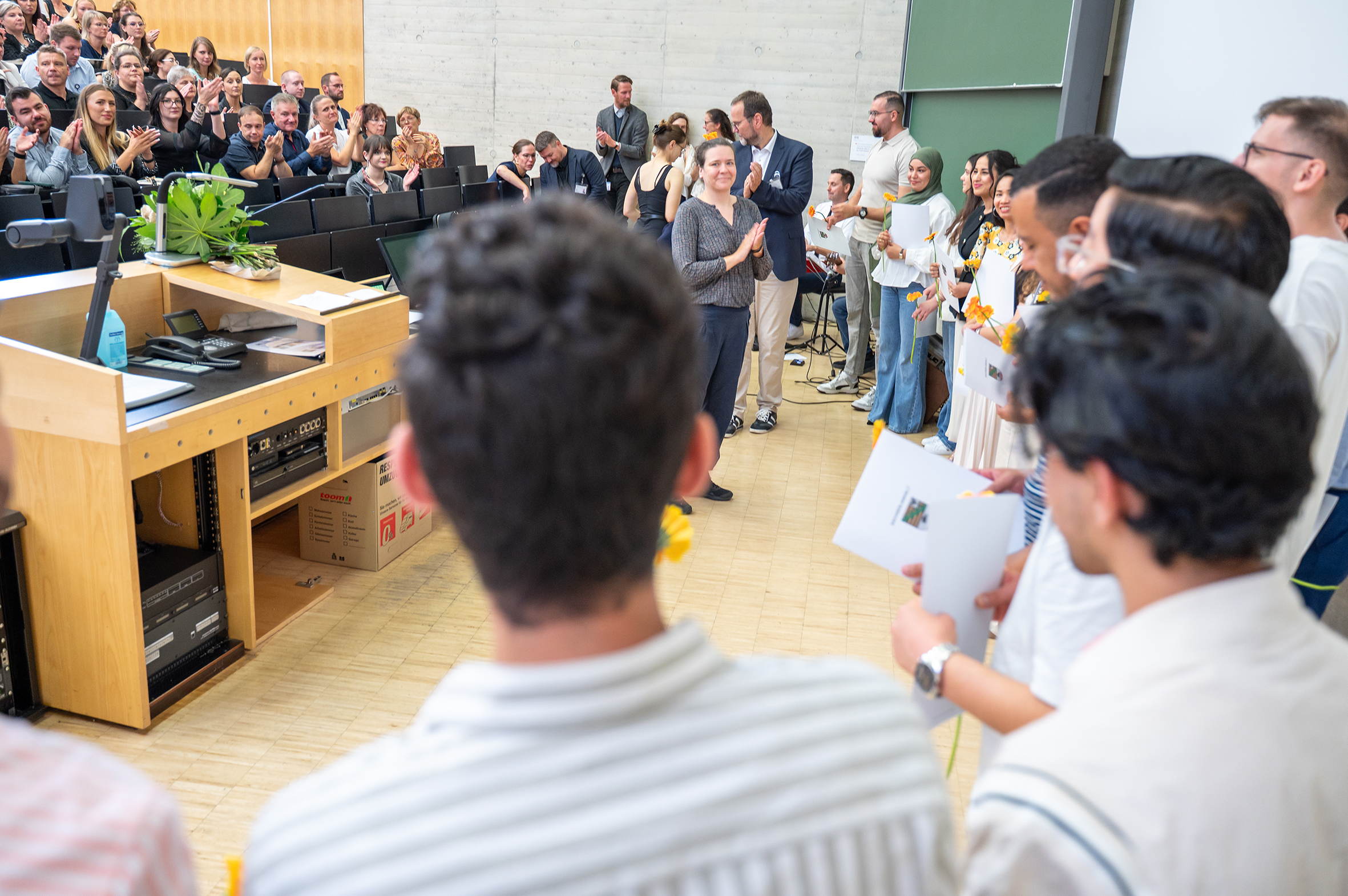 Eine Gruppe von Menschen steht in einem Hörsaal. Eine Person verteilt Abschlusszeugnisse an die Gruppe. Im Vordergrund und im Hintergrund befinden sich weitere Personen. Auch in den Sitzreihen des Hörsaals sitzen Personen.