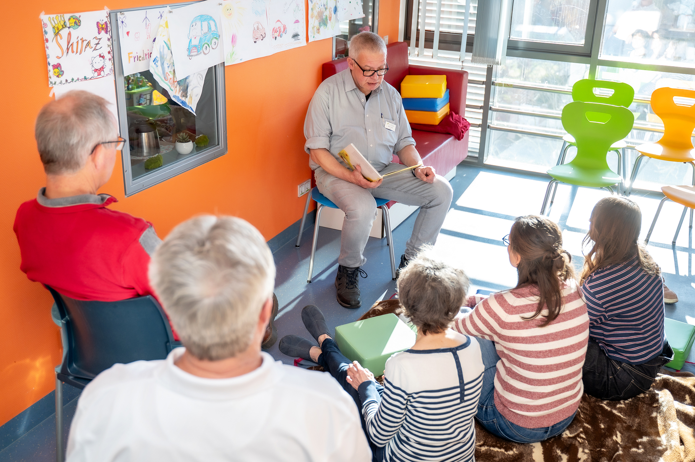 Lesung in einem Aufenthaltsraum einer Kinderstation eines Krankenhauses. Ein Mann sitzt auf einem Stuhl und liest vor. Vor ihm sitzen mehrere Personen auf dem Boden und auf Stühlen und hören zu.