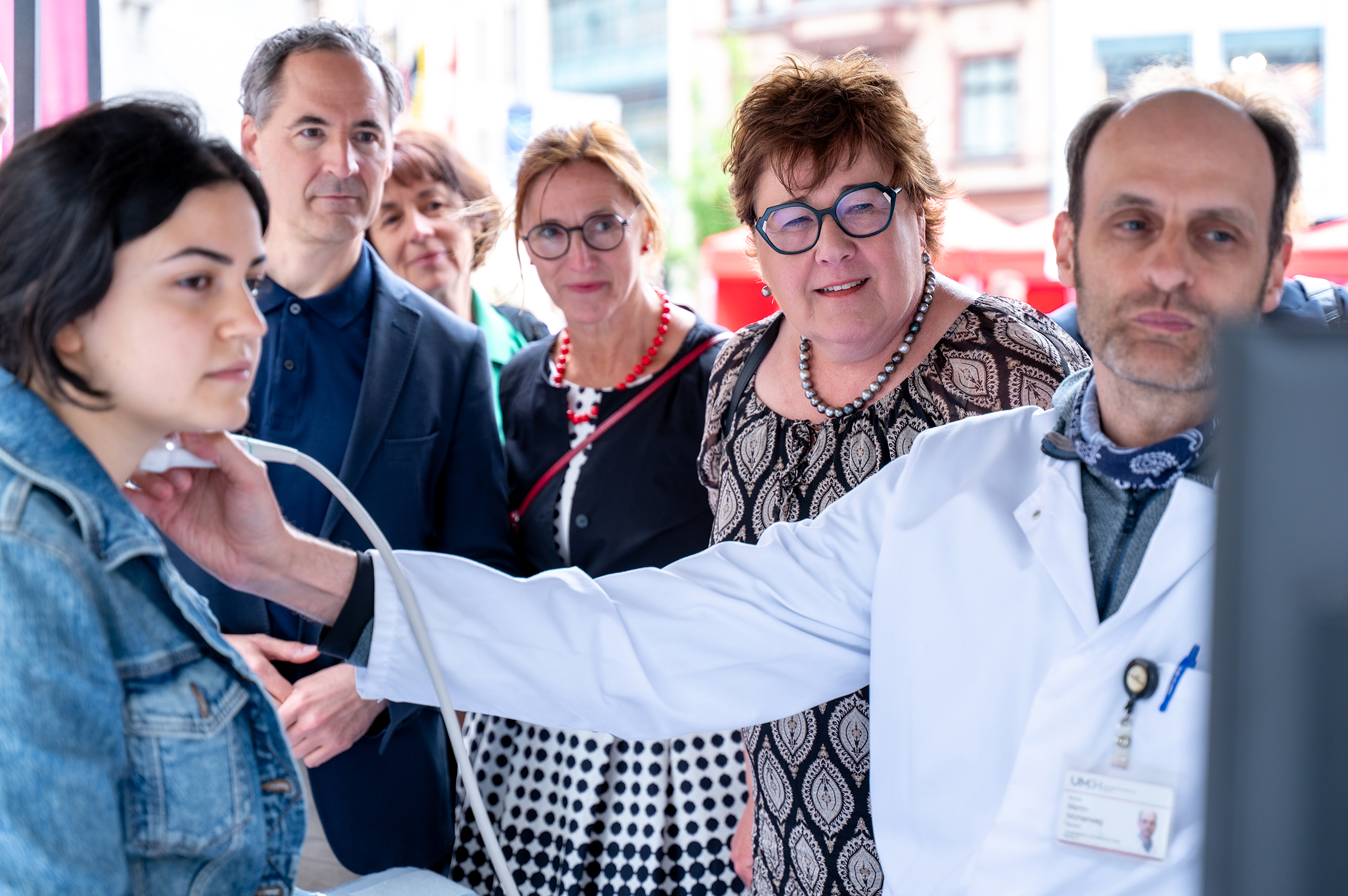 Eine Person in medizinischer Arbeitskleidung führt mithilfe eines medizinischen Geräts eine Untersuchung am Hals einer anderen Person durch. Im Hintergrund stehen drei weitere Personen. Alle schauen auf einen Bildschirm.
