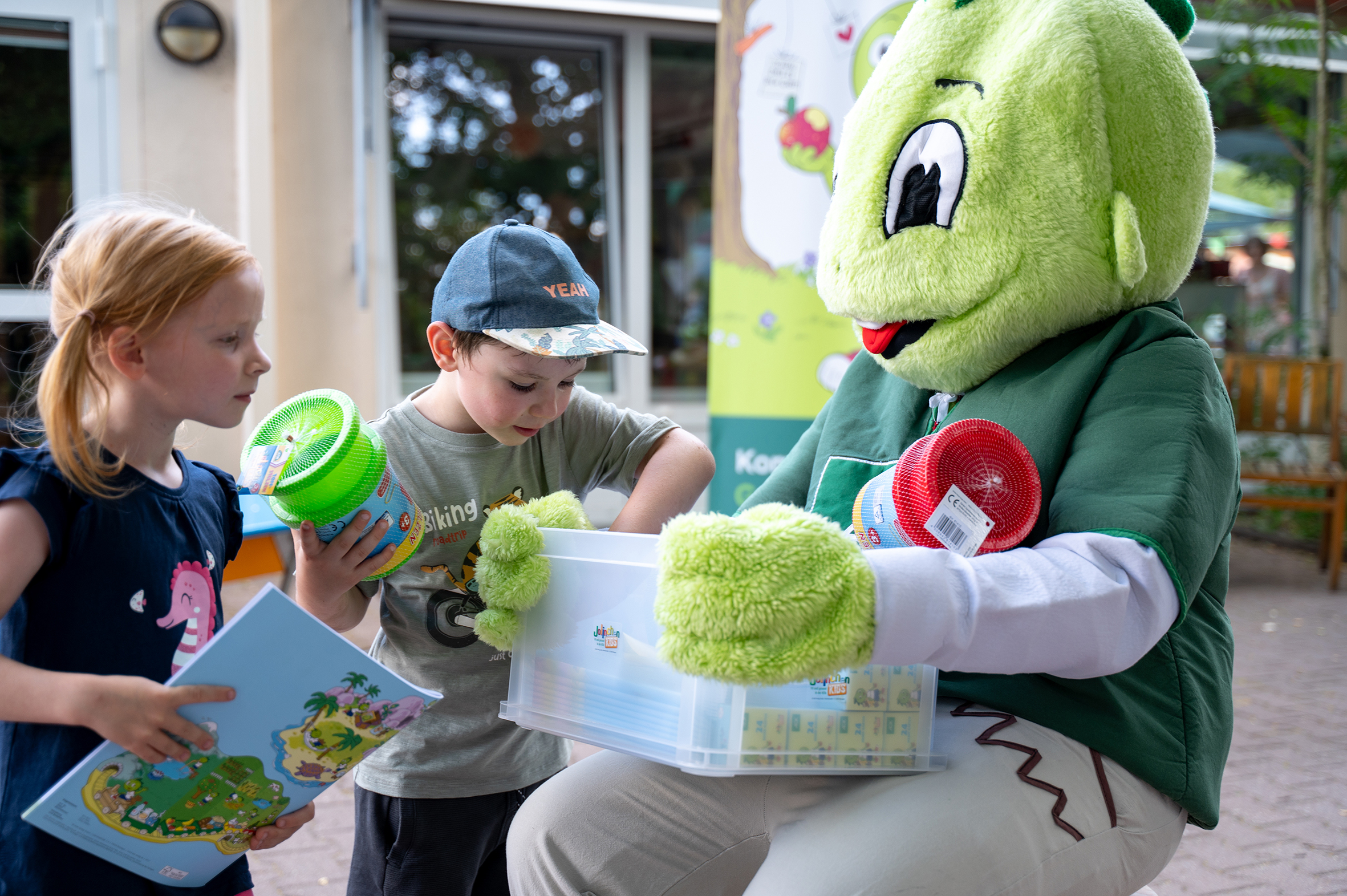 Das AOK-Maskottchen Jolinchen kniet auf dem Außengelände der Kita des UKH auf dem Boden und hält eine Kiste in den Händen. Zwei Kinder suchen sich Spielsachen aus der Kiste heraus.