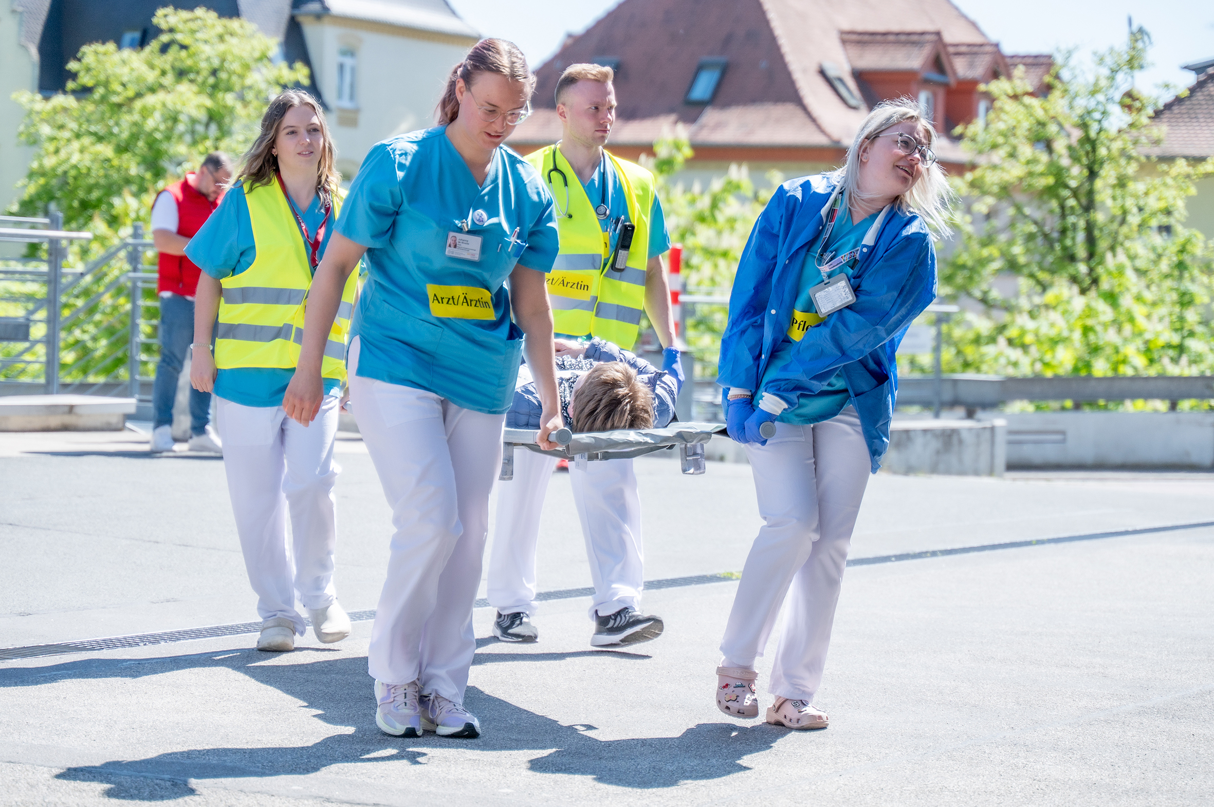 Vier Personen in medizinischer Berufskleidung tragen eine Person auf einer Trage vom Vorplatz der Notaufnahme ins Gebäude. Die zwei hinteren Personen tragen Warnwesten über ihrer Kleidung.