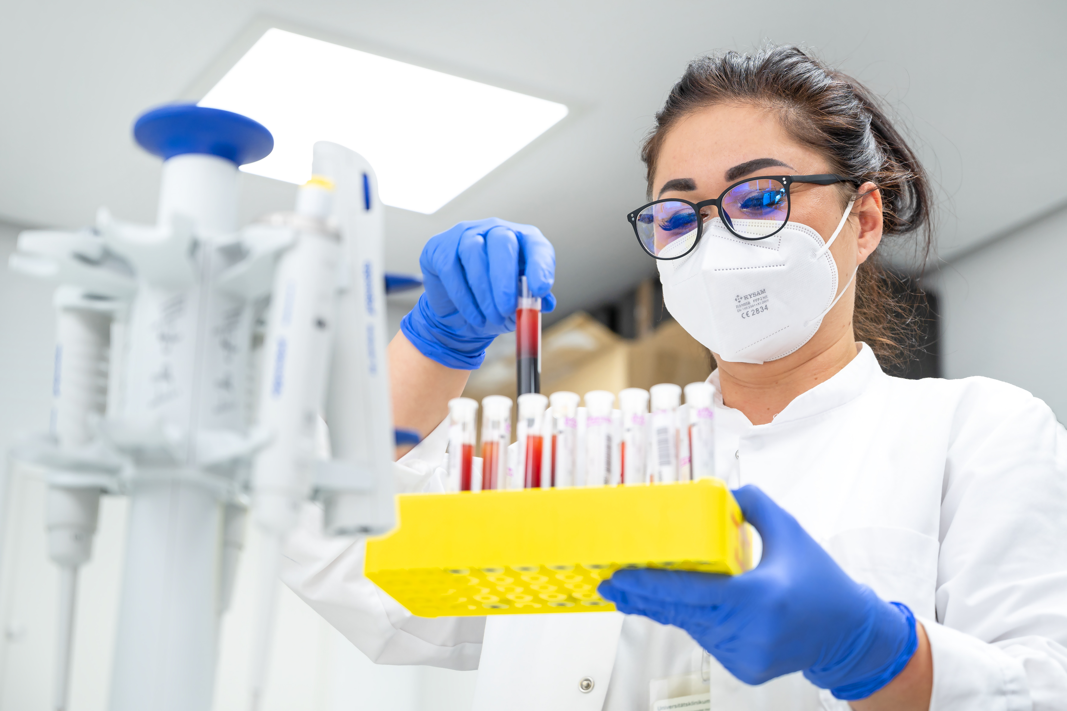 Eine Frau mit Brille, FFP2-Maske und medizinischen Einmalhandschuhen hält in der linken Hand ein Rack voller Röhrchen. Die Gefäße sind mit einer roten Flüssigkeit gefüllt. Mit der rechten Hand entnimmt sie eines davon. Im Vordergrund ist unscharf ein Pipettenständer erkennbar. Der Raum ist lichtdurchflutet.