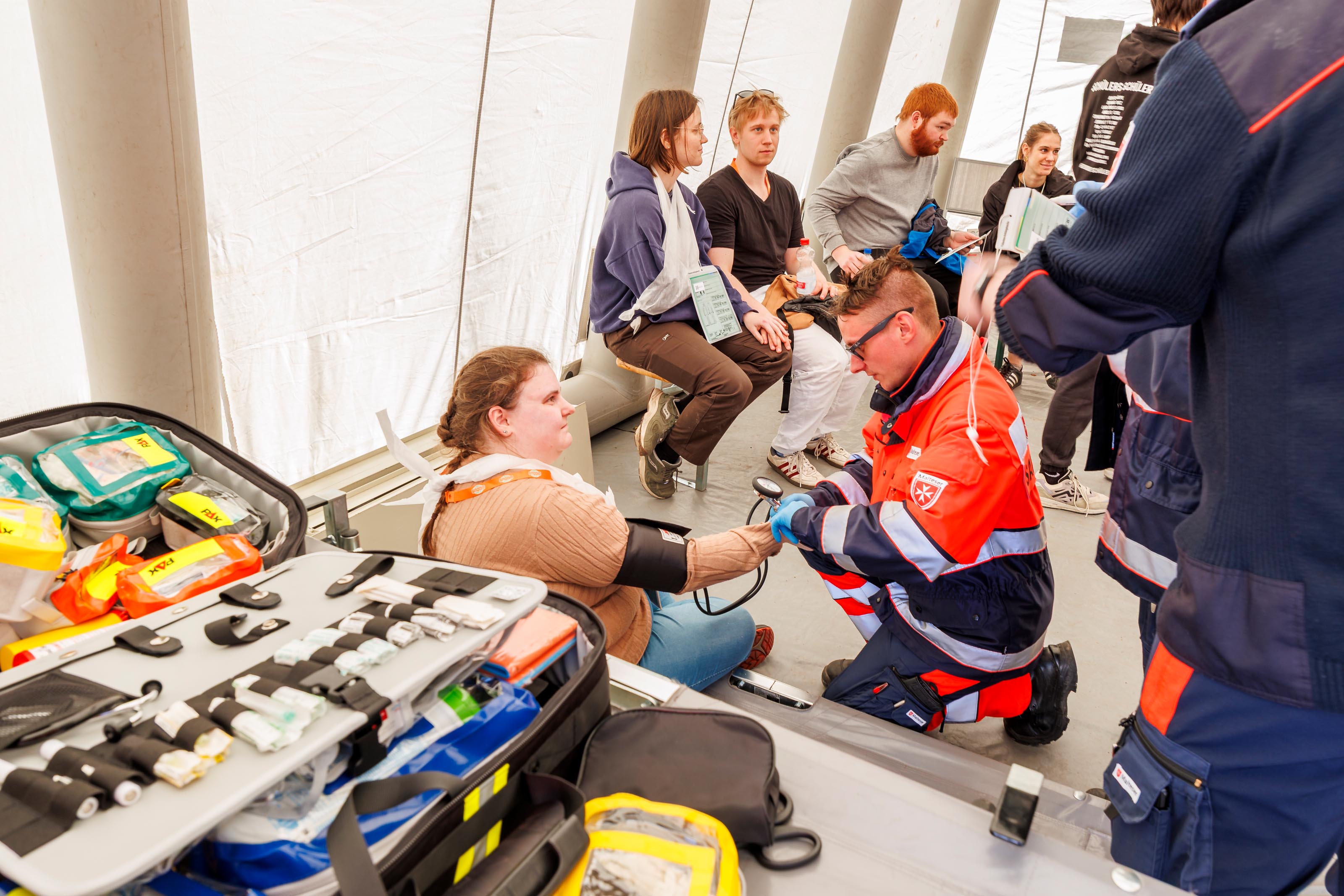 Eine Person in Sanitäter- oder Notarztkleidung versorgt eine Patientin. Im Hintergrund sitzen weitere Personen. Im Vordergrund liegt ein aufgeklappter Notarztkoffer.