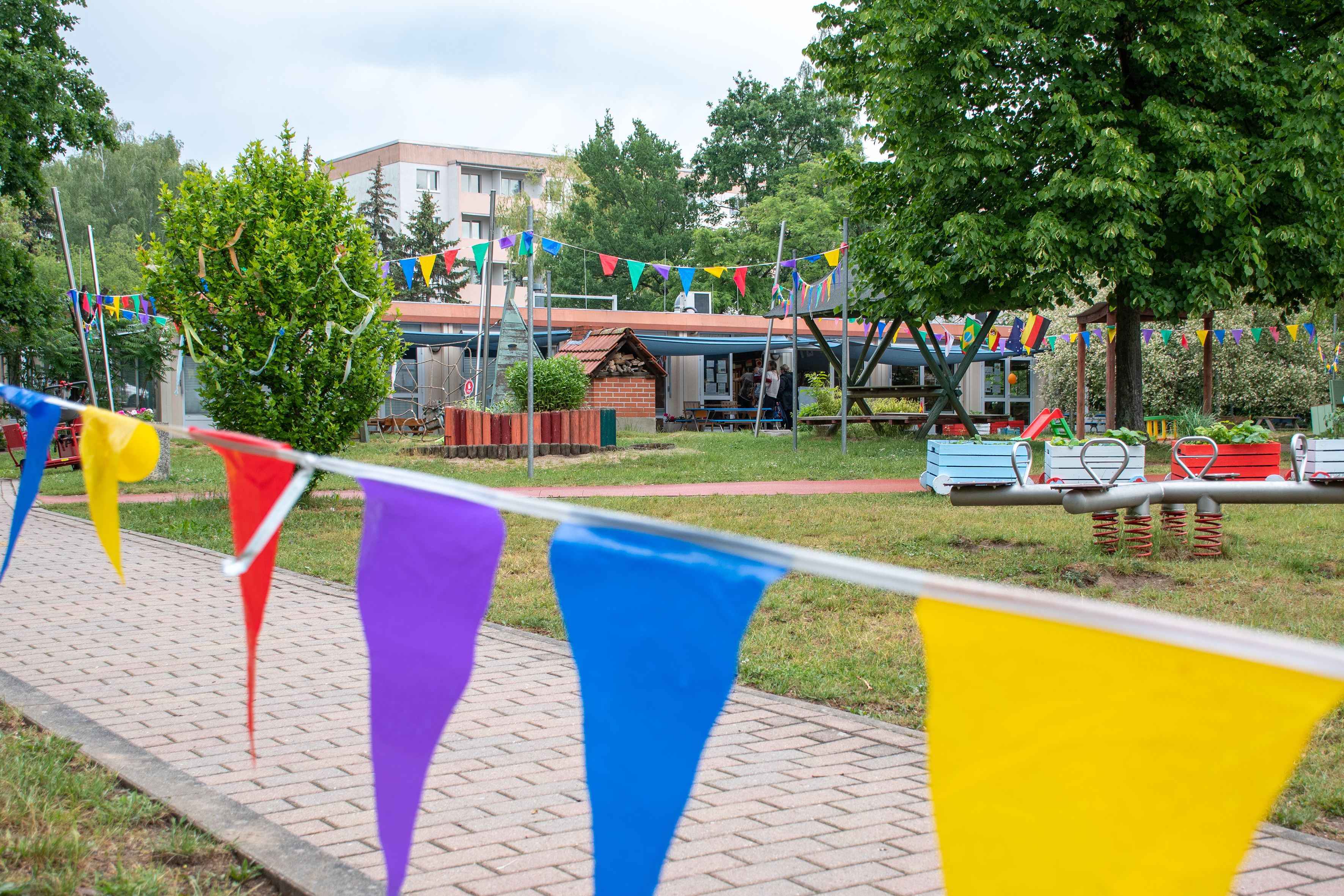 Außenbereich der Kita MediKids. Auf der Wiese stehen Spielsachen, im Hintergrund befindet sich das Hauptgebäude der Kita. Im Vordergrund hängt eine bunte Wimpelgirlande.