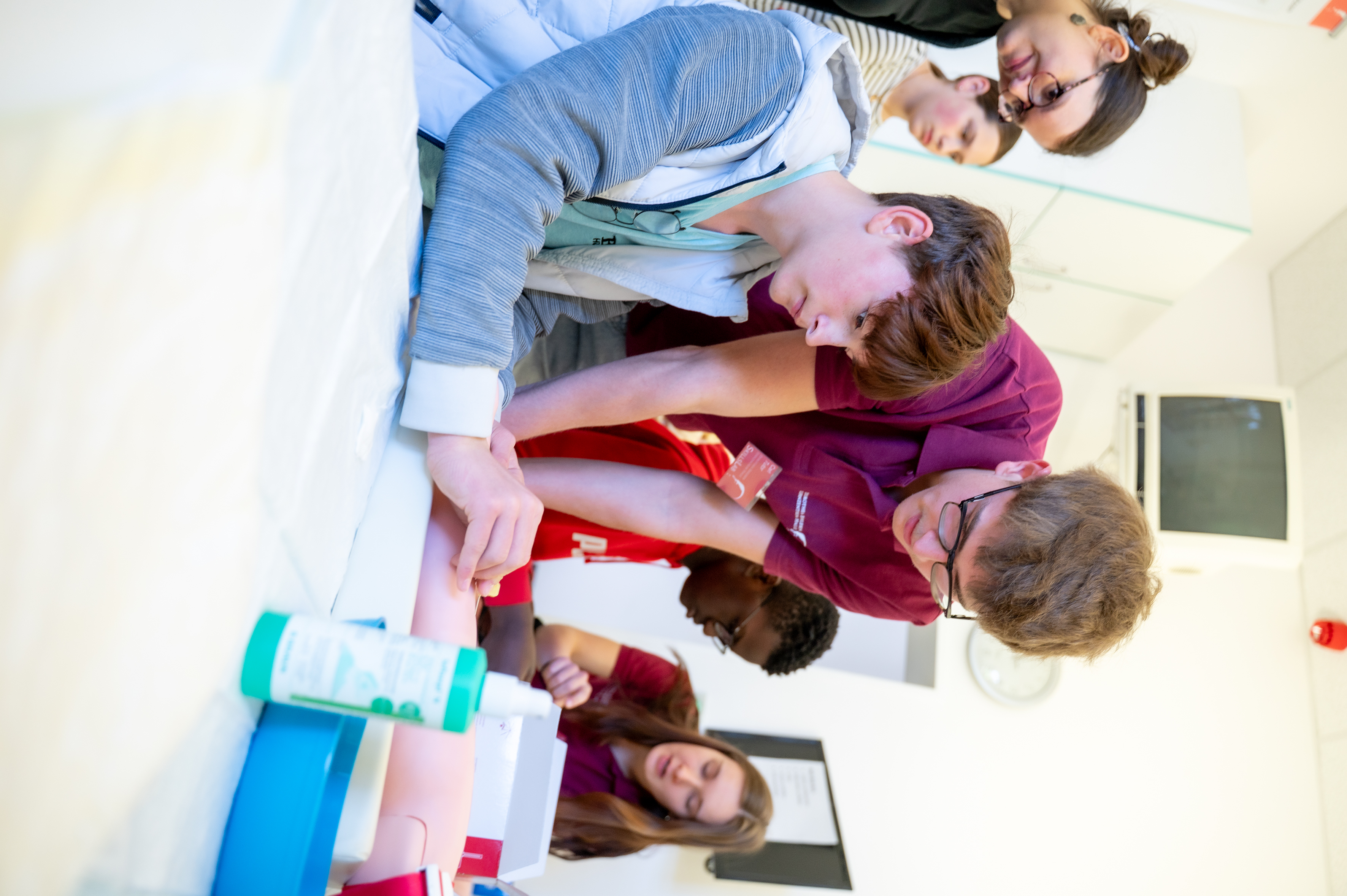 Eine Person zeigt einem Schüler, wie man Blut abnimmt. Im Hintergrund befinden sich weitere Schüler und andere Personen. Rechts auf dem Tisch, an dem der Schüler sitzt, stehen medizinische Materialien.