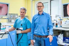 Dr. Markus Reitmann und Prof. Dr. Roland Haase stehen nebeneinander in einem medizinischen Behandlungszimmer. Dr. Reitmann hält ein Endoskopiergerät in den Händen, ein langer schwarzer Schlauch, an dessen Spitze ein Licht leuchtet. Auf Monitoren ist das Innere einer Speiseröhre zu sehen. Dr. Reitmann trägt blaue medizinische Arbeitskleidung, Prof. Haase ein blaues Hemd und Jeanshose.