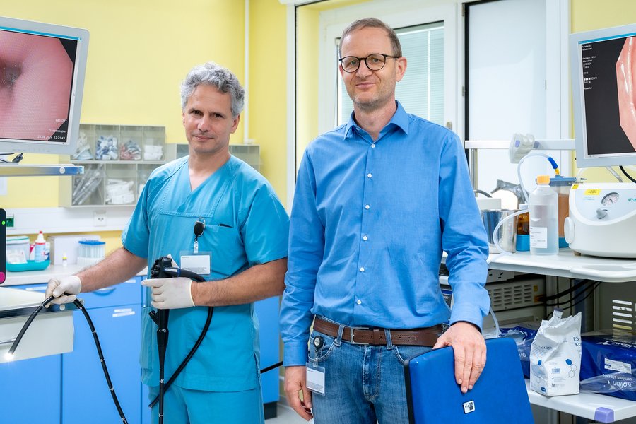 Dr. Markus Reitmann und Prof. Dr. Roland Haase stehen nebeneinander in einem medizinischen Behandlungszimmer. Dr. Reitmann hält ein Endoskopiergerät in den Händen, ein langer schwarzer Schlauch, an dessen Spitze ein Licht leuchtet. Auf Monitoren ist das Innere einer Speiseröhre zu sehen. Dr. Reitmann trägt blaue medizinische Arbeitskleidung, Prof. Haase ein blaues Hemd und Jeanshose.  Dr. Markus Reitmann und Prof. Dr. Roland Haase stehen nebeneinander in einem medizinischen Behandlungszimmer. Dr. Reitmann hält ein Endoskopiergerät in den Händen, ein langer schwarzer Schlauch, an dessen Spitze ein Licht leuchtet. Auf Monitoren ist das Innere einer Speiseröhre zu sehen. Dr. Reitmann trägt blaue medizinische Arbeitskleidung, Prof. Haase ein blaues Hemd und Jeanshose.