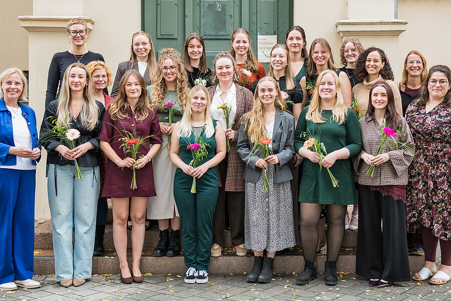 Auf dem Gruppenfoto sind 21 Personen zu sehen. Die 16 anwesenden Absolventinnen des Studiengangs Hebammenwissenschaft halten jeweils einen kleinen Blumenstrauß in den Händen. Auf dem Bild sind außerdem Lehrende und die Studiengangsleitung zu sehen. Auf dem Gruppenfoto sind 21 Personen zu sehen. Die 16 anwesenden Absolventinnen des Studiengangs Hebammenwissenschaft halten jeweils einen kleinen Blumenstrauß in den Händen. Auf dem Bild sind außerdem Lehrende und die Studiengangsleitung zu sehen.
