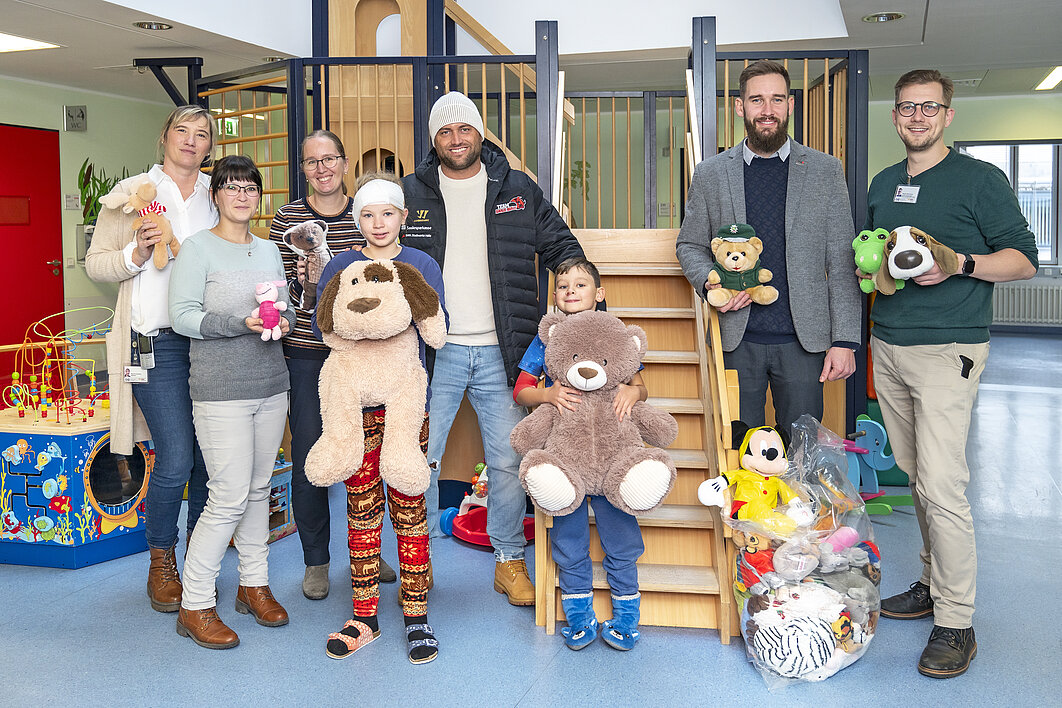 Gruppenfoto vor einem Spielplatz auf einer Kinder-Station im Krankenhaus. Die sechs Erwachsenen und zwei Kinder halten Kuscheltiere in den Händen und gucken in die Kamera.