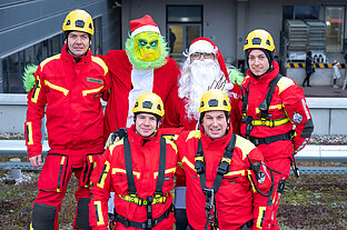 Gruppenfoto mit vier Männern in Höhenretter-Uniform mit Helmen und zwei Männern, die als Grinch und als Weihnachtsmann verkleidet sind.