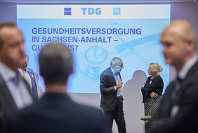 Im Vordergrund sind drei Personen unscharf zu erkennen. Im Hintergrund unterhalten sich drei weitere Personen. Die Personen im Hintergrund stehen in der Projektionsfläche eines Beamers. An der Wand sind der Text „Gesundheitsversorgung in Sachsen-Anhalt - Quo vadis?“ und verschiedene Logos zu sehen.