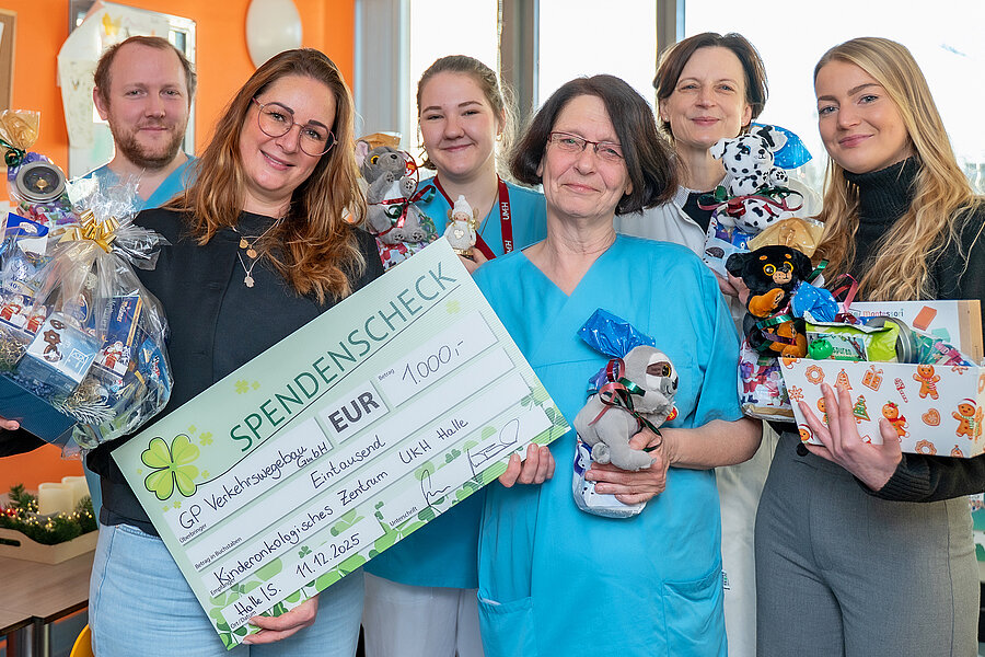 Sechs Personen stehen im Aufenthaltsraum einer Kinderstation im UKH. Einige Personen halten einen Spendenscheck über 1000 Euro und Geschenke in den Händen. Sechs Personen stehen im Aufenthaltsraum einer Kinderstation im UKH. Einige Personen halten einen Spendenscheck über 1000 Euro und Geschenke in den Händen.