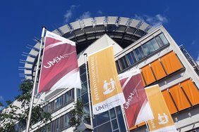 Foto des Hauptgebäudes des Universitätsklinikums Halle (Saale) aus der Froschperspektive. Im Vordergrund befinden sich vier Fahnenmäste. Die erste und die dritte Fahne von links zeigen das UMH-Logo und die Corporate-Design-Farbe. Auf der zweiten und vierten Fahne von links steht "Patientensicherheit" und "World Patient Safety Day". Unten befindet sich das UMH-Logo, rechts darüber sind zwei Figuren schemenhaft abgebildet.