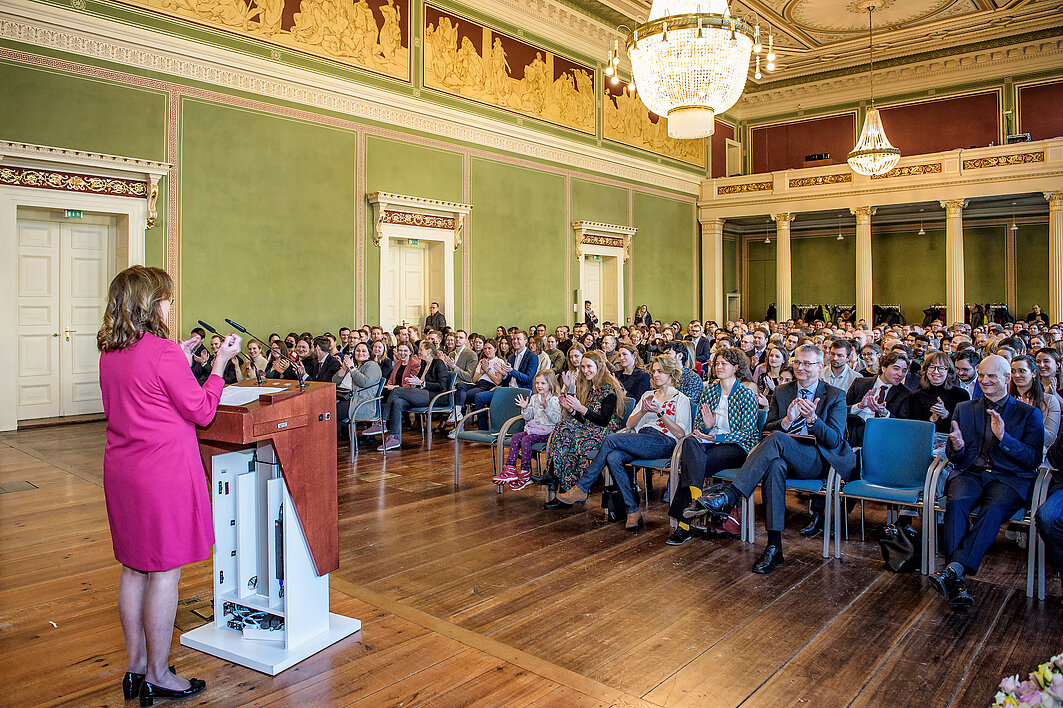 Die Dekanin steht an einem Pult in der Aula des Löwengebäudes. Der Raum ist voller Menschen.