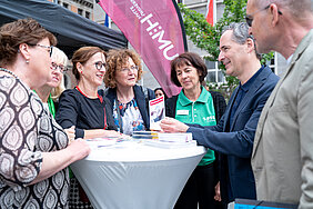 Sieben Personen stehen auf dem Marktplatz in Halle (Saale) um einen Stehtisch herum und unterhalten sich. Im Hintergrund befindet sich eine Beachflag der Universitätsmedizin Halle.