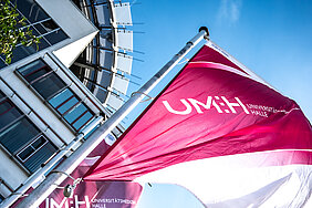 magentafarbene Fahne mit Aufdruck UMH flattert vor blauem Himmel und einer angeschnittenen Fassade im Wind.