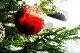 Nahaufnahme einer großen roten Christbaumkugel, die an einem Weihnachtsbaum hängt. Im Hintergrund ist das Hauptgebäude des Universitätsklinikums Halle (Saale) zu sehen.