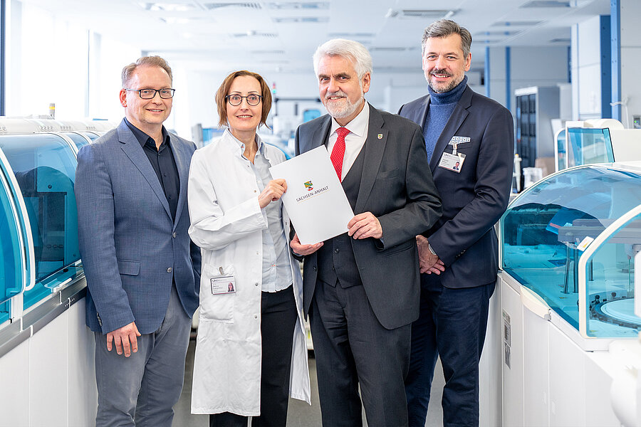 drei Männer und eine Frau stehen in einem Labor mit Laborgeräten im Hintergrund. Es wird ein Fördermittelbescheid übergeben. drei Männer und eine Frau stehen in einem Labor mit Laborgeräten im Hintergrund. Es wird ein Fördermittelbescheid übergeben.