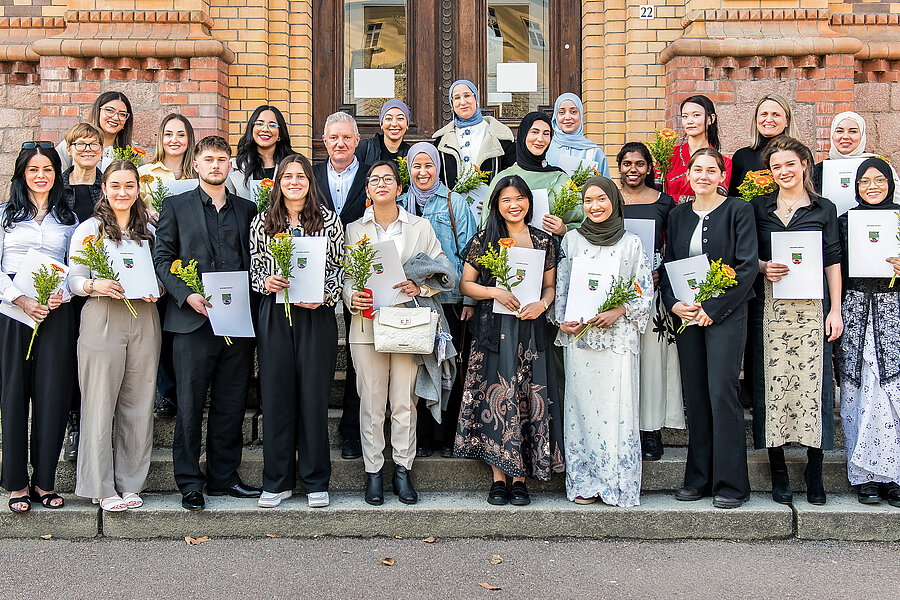 Die Absolvent:innen stehen vor einem Gebäude und schauen in die Kamera. Sie halten ihre Zeugnisse und Blumen in den Händen. Die Absolvent:innen stehen vor einem Gebäude und schauen in die Kamera. Sie halten ihre Zeugnisse und Blumen in den Händen.