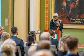 Eine Frau mit weißen Haaren und einer runden schwarzen Brille steht an einem Pult mit zwei Mikrofonen und hält einen Vortrag. Dabei gestikuliert sie mit ihrer linken Hand, in der sie ein Präsentationsgerät hält.