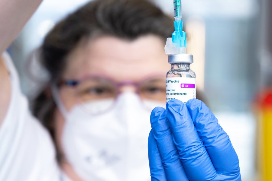 Im Hintergrund ist unscharf das Gesicht einer Person mit Mund-Nasen-Schutz und Brille zu erkennen, die auf eine Impfstoffflasche in ihrer Hand schaut, die sich im Bildfokus befindet. Sie hält das Fläschchen mit einem blauen medizinischen Nitrilhandschuh in der linken Hand und zieht mit der rechten Hand eine Dosis aus einer Spritze auf. Auf dem Fläschchen ist u. a. das Wort „Vaccine“ zu erkennen.  Im Hintergrund ist unscharf das Gesicht einer Person mit Mund-Nasen-Schutz und Brille zu erkennen, die auf eine Impfstoffflasche in ihrer Hand schaut, die sich im Bildfokus befindet. Sie hält das Fläschchen mit einem blauen medizinischen Nitrilhandschuh in der linken Hand und zieht mit der rechten Hand eine Dosis aus einer Spritze auf. Auf dem Fläschchen ist u. a. das Wort „Vaccine“ zu erkennen.