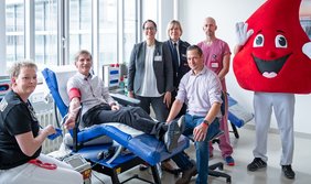 Gruppenbild beim Blutspendedienst: Ein Mann liegt auf einer blauen Liege, er hält den rechten Arm nach unten ausgestreckt, daran hängt eine Blutspendenadel. Neben ihm stehen und sitzen drei Frauen und zwei Männer sowie ein Mensch im Maskottchenkostüm eines freundlich schauen-den Blutstropfens. Eine Frau trägt eine Polizeiuniform, ein Mann beerenfarbene medizinische Arbeitskleidung. Die anderen sind in zivil gekleidet.