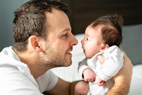 Ein junger Vater hält sein neugeborenes Baby fest. Sie schauen sich an, der Vater lächelt, das Baby hat die Augen weit geöffnet. Beide sind im Profil zu sehen, die Nasenspitzen berühren sich beinahe.
