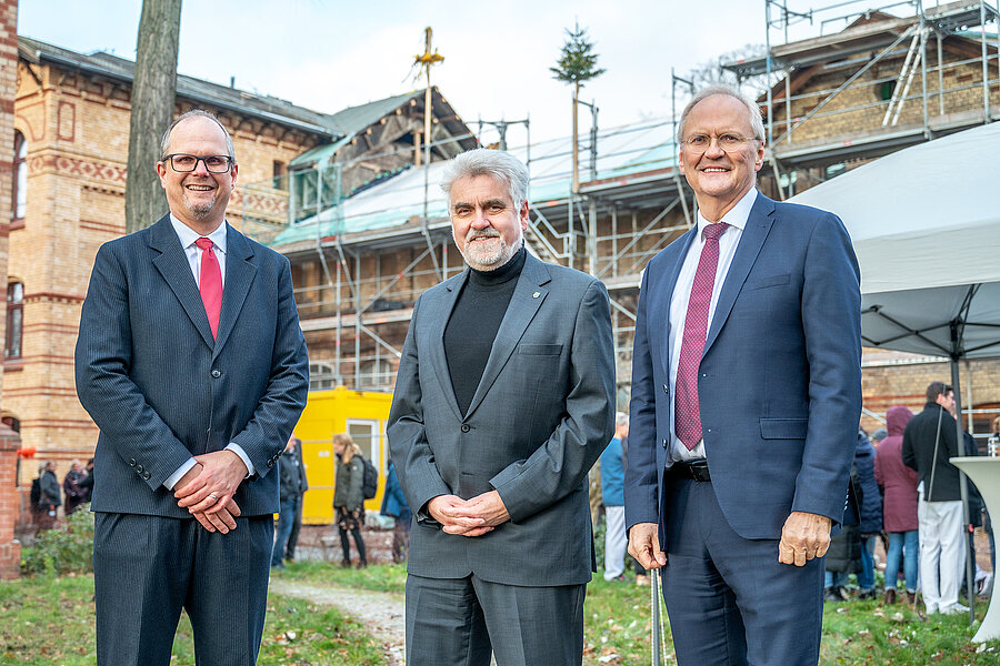 Drei Männer im Anzug stehen vor einem Backsteingebäude, das eingerüstet ist. Auf dem Dachstuhl ist unscharf ein Richtkranz zu erkennen.  Drei Männer im Anzug stehen vor einem Backsteingebäude, das eingerüstet ist. Auf dem Dachstuhl ist unscharf ein Richtkranz zu erkennen.