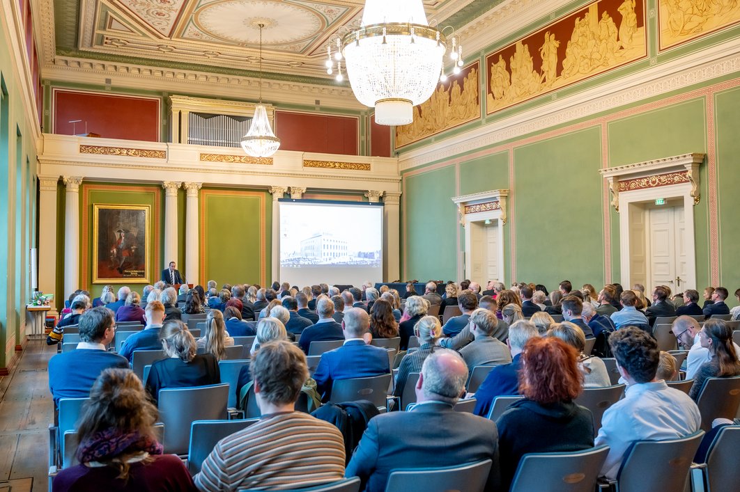 Die historische Aula im Löwengebäude der MLU ist imposant geschmückt und von mehreren großen Kronleuchtern erhellt. Zum Akademischen Festakt haben sich wieder hunderte Menschen im Festsaal versammelt. Auf einer Projektionsfläche ist das Löwengebäude im 19. Jahrhundert zu sehen.