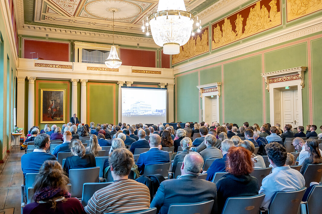 Die historische Aula im Löwengebäude der MLU ist imposant geschmückt und von mehreren großen Kronleuchtern erhellt. Zum Akademischen Festakt haben sich wieder hunderte Menschen im Festsaal versammelt. Auf einer Projektionsfläche ist das Löwengebäude im 19. Jahrhundert zu sehen.