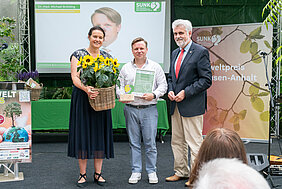 Drei Personen stehen auf einer Bühne und schauen in die Kamera. Die Person links hält einen Strauß Sonnenblumen in den Händen. Die Preson in der Mitte hält eine Urkunde und den Umwelt-Ehrenpreis in den Händen.