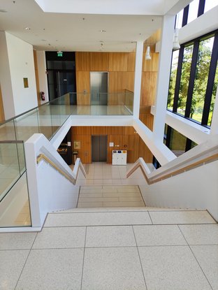 Eindruck von einem Treppenhaus, der Boden ist mit hellen großen Fliesen oder Steinplatten belegt. Die Treppe ist von weißen halbhohen Wänden eingefasst, auf der Etage selbst bildet eine Glasbalustrade die Grenze. Rechts ist eine Fensterfront zu sehen.