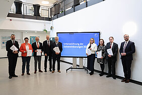Neun Personen stehen in einer Reihe nebeneinander im Raum des Forschungscampus STIMULATE. In der Mitte der Reihe befindet sich ein großer Bildschirm mit dem Bildtext „Unterzeichnung der Zielvereinbarung“.