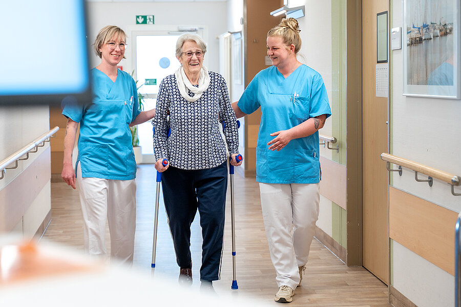 Zwei Personen in medizinischer Arbeitskleidung begleiten eine ältere Person in einem Krankenhausflur. Die ältere Person geht in der Mitte an Krücken und wird von den beiden Personen leicht gestützt. Zwei Personen in medizinischer Arbeitskleidung begleiten eine ältere Person in einem Krankenhausflur. Die ältere Person geht in der Mitte an Krücken und wird von den beiden Personen leicht gestützt.