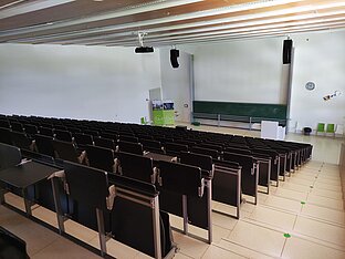 Einer der beiden Hörsäle des BMC, charakterisiert durch getreppte dunkle Reihen mit Klappsitzen und weißen Wänden. Im Hintergrund ist eine große grüne Tafel angebracht, an der Decke hängen Lautsprecher und Beamer.