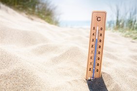 Ein Thermometer steckt im Sand und zeigt 35°C an. Im Hintergrund befinden sich Gräser und das Meer. Der Fokus liegt auf dem Thermometer, der Hintergrund ist unscharf.  