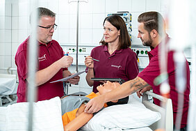 Drei Personen stehen in einem Raum des DELH. Im Vordergrund liegt eine Simulationspuppe in einem Bett. Eine Person berührt den Simulator an der Schulter, die anderen beiden Personen halten Stifte und ein Tablet bzw. ein Dokument in den Händen.