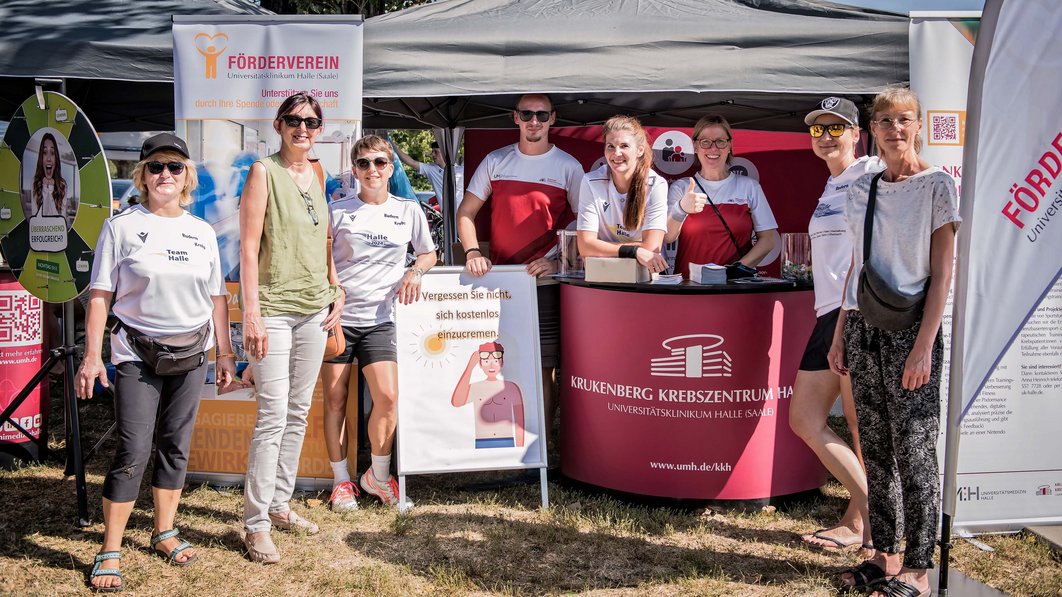 Gruppenbild. Einige Personen stehen vor einem Infostand des Fördervereins des Universitätsklinikums Halle (Saale) und des Krukenberg-Krebszentrums Halle, einige dahinter. Vor dem Infostand steht ein Aufsteller, dahinter ein Banner. Auf dem Tresen befindet sich Infomaterial.