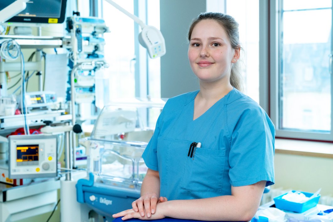 Eine junge Frau in blauer medizinischer Arbeitskleidung. Im Hintergrund sieht man medizinische Geräte einer Intensivstation sowie einen Inkubator für Neugeborene.