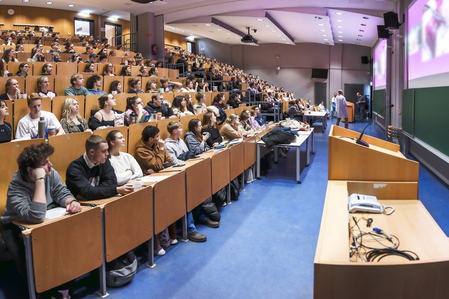 Ein großer Hörsaal voller Studierender, die auf eine große Projektion blicken, auf der verschwommen eine Operation zu sehen ist.  Ein großer Hörsaal voller Studierender, die auf eine große Projektion blicken, auf der verschwommen eine Operation zu sehen ist.