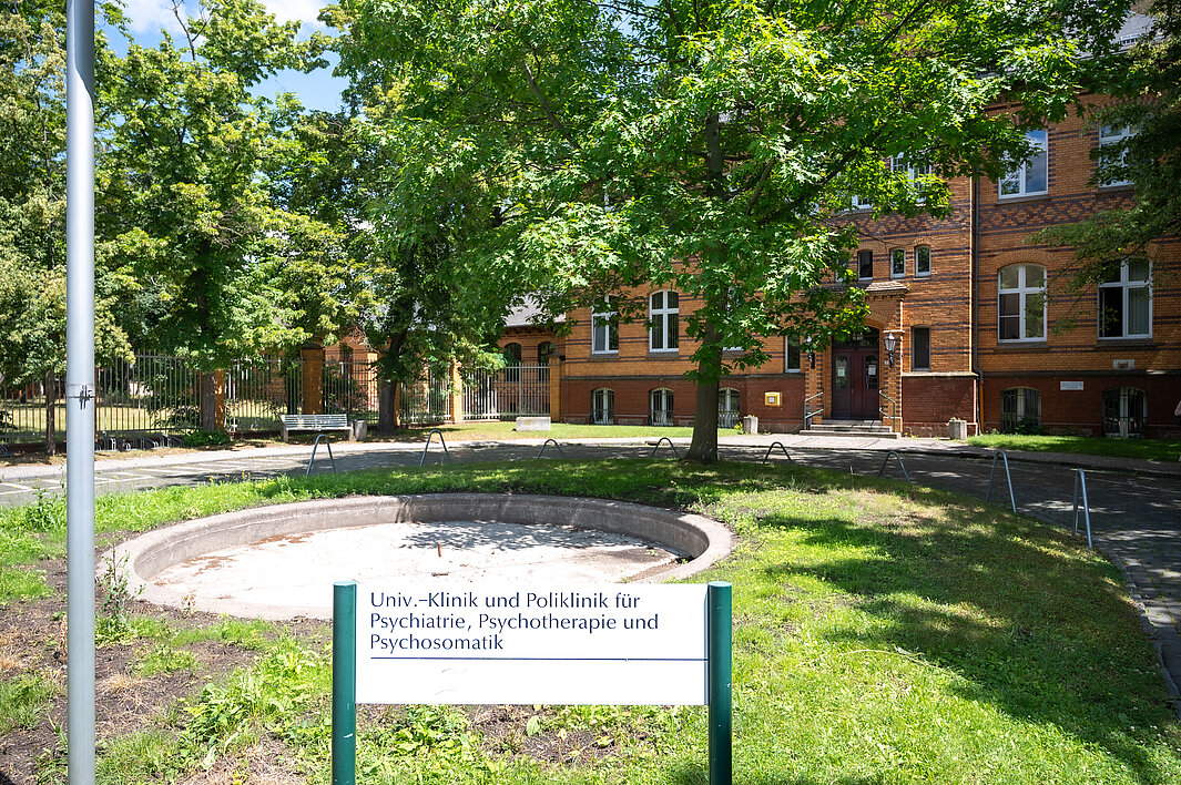 Außenansicht des Haupteingangs der Psychiatrischen Klinik des UKH. Im Vordergrund befindet sich ein Schild mit dem Kliniknamen. Dahinter befindet sich der ehemalige Springbrunnen. Im Hintergrund steht das Eingangsgebäude.