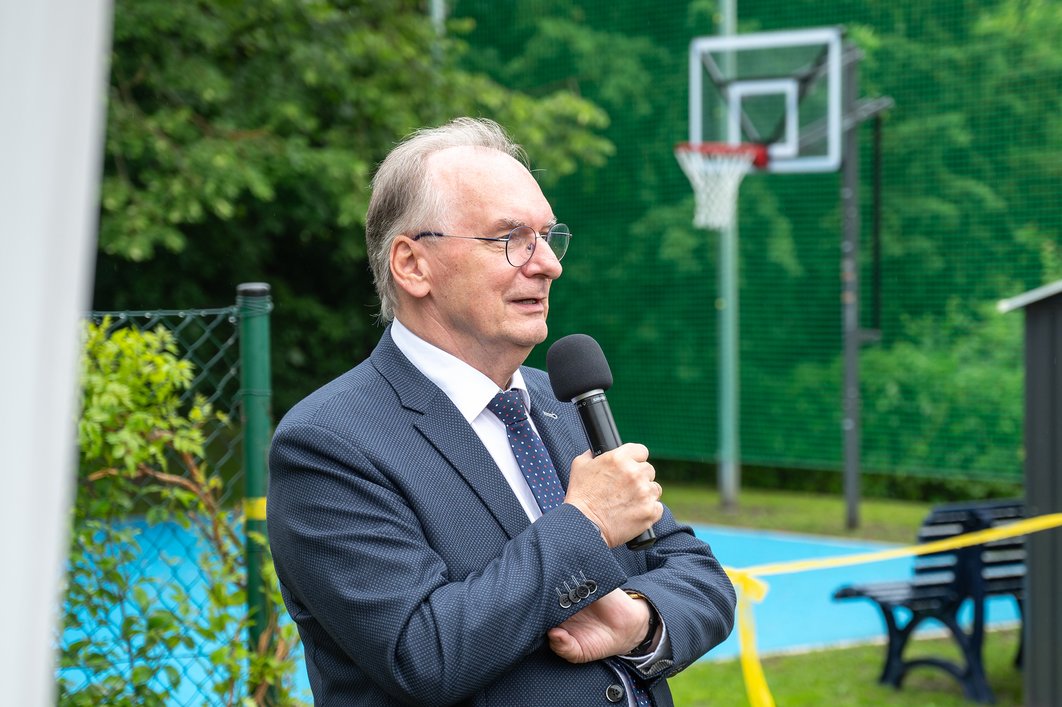 Mann mit grauen Haaren und Brille hält Mikro in der Hand. Im Hintergrund ist ein Basketballplatz zu sehen.