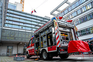 Blick aus einem Hof von schräg unten hoch zu Gebäuden mit Glasfassaden. Im Vordergrund steht ein Feuerwehrauto mit geöffneten Gerätelagern. Ganz oben sind in klein zwei als Grinch und als Weihnachtsmann verkleidete Personen an einem Sicherungsseil zu sehen, die sich abseilen.